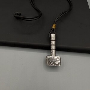Puede incluir: Un collar con colgante plateado con un dise&ntilde;o de martillo. La cabeza del martillo tiene una superficie estampada y el mango es cil&iacute;ndrico con l&iacute;neas horizontales. El collar tiene un cord&oacute;n negro.