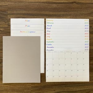 May include: A set of white paper planner sheets with rainbow-colored text. The sheets include sections for ideas, plans, income & expenses, and a 2027 calendar. A gray card is on the left.