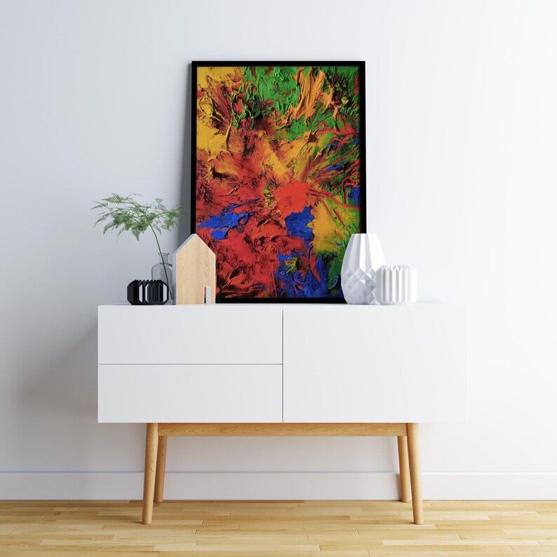 K&ouml;nnte beinhalten: Ein wei&szlig;er Schrank mit zwei Schubladen und Holzbeinen, auf dem ein gerahmtes abstraktes Gem&auml;lde mit leuchtenden Farben in Rot, Gelb, Blau und Gr&uuml;n h&auml;ngt. Das Gem&auml;lde befindet sich an einer wei&szlig;en Wand mit einem kleinen Holzhaus und einer Pflanze in einer Glasvase auf dem Schrank.