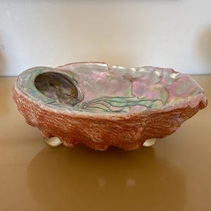May include: A large, iridescent seashell bowl with a pink, green, and white interior. The bowl is resting on three small, round feet.