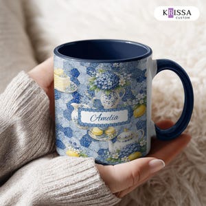 May include: A ceramic mug with a dark blue interior and handle, featuring a white rectangular label with the name Amelia printed in blue script. The exterior surface displays a blue and white mosaic pattern with illustrations of lemons, flowers, and tea sets.