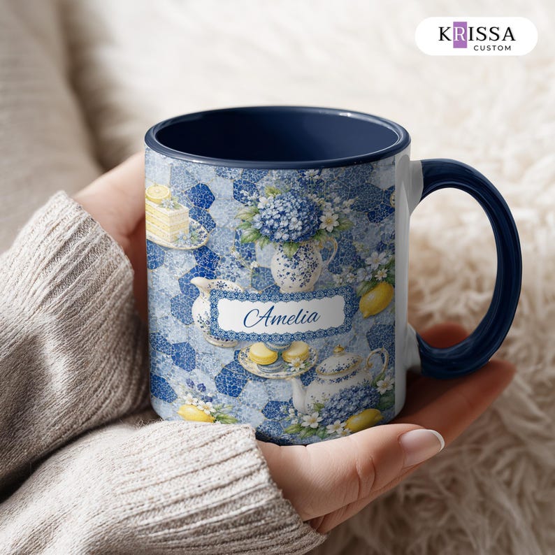 May include: A ceramic mug with a dark blue interior and handle, featuring a white rectangular label with the name Amelia printed in blue script. The exterior surface displays a blue and white mosaic pattern with illustrations of lemons, flowers, and tea sets.