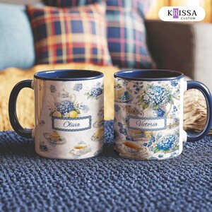 May include: Two ceramic mugs with dark blue handles and rims, featuring a white base with a blue and white floral pattern of hydrangeas, teapots, and teacups. One mug displays the name Olivia and the other displays the name Victoria in script.
