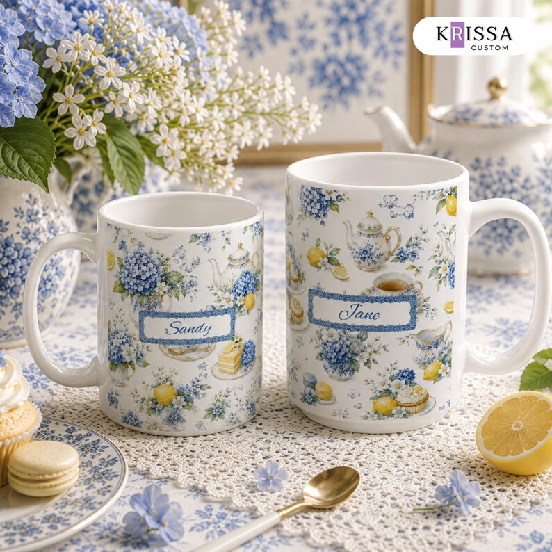 May include: Two white ceramic mugs featuring a blue and yellow floral pattern with illustrations of teapots, lemons, and cakes. One mug is personalized with the name Sandy, and the other with the name Jane, both inside blue rectangular frames.