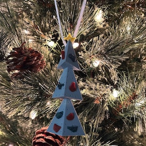 May include: A light blue Christmas tree ornament with colorful climbing holds. The ornament is shaped like a tree and has a yellow star at the top. It hangs from a white ribbon. The ornament is hanging on a Christmas tree.