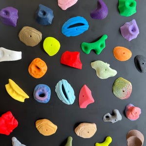 May include: A collection of colorful climbing holds in various shapes and sizes, including rounded, curved, and textured designs. The holds are in shades of pink, blue, green, yellow, orange, and brown, arranged on a black surface.