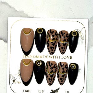 May include: A set of almond-shaped press-on nails. The nails feature a variety of designs, including solid black, tan with gold accents, and leopard print with gold stripes. The card reads "HANDMADE WITH LOVE" and includes size labels.