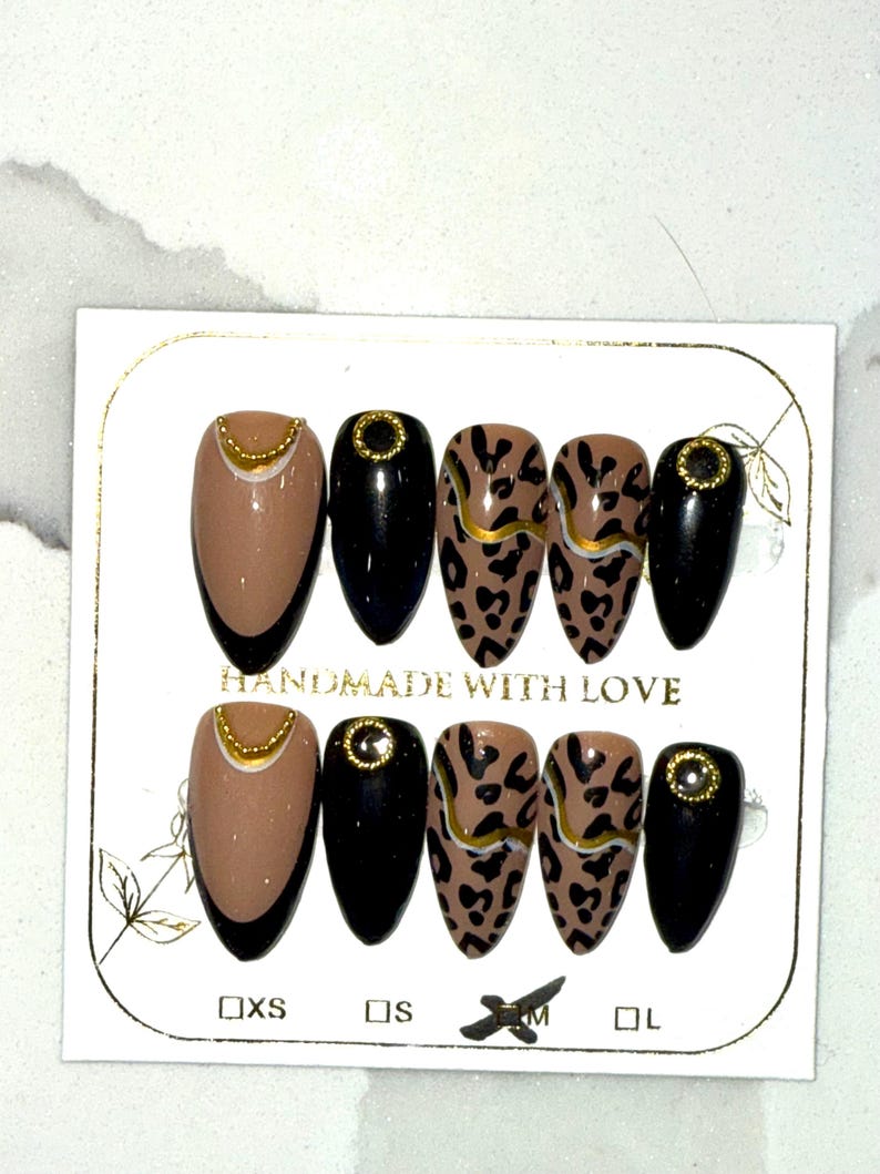 May include: A set of almond-shaped press-on nails. The nails feature a variety of designs, including solid black, tan with gold accents, and leopard print with gold stripes. The card reads "HANDMADE WITH LOVE" and includes size labels.