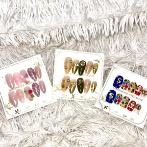 May include: Three sets of artificial nails on display. One set features pink and glitter nails with floral accents. Another set has gold and olive green nails with gold embellishments. The third set has colorful designs.