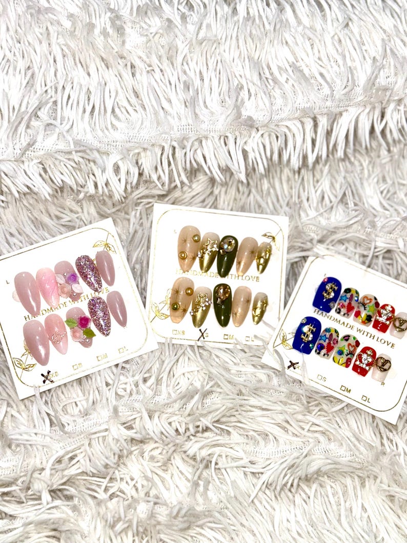 May include: Three sets of artificial nails on display. One set features pink and glitter nails with floral accents. Another set has gold and olive green nails with gold embellishments. The third set has colorful designs.