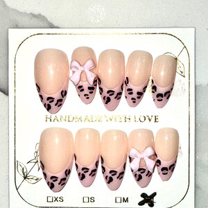 May include: A set of almond-shaped press-on nails. The nails are a nude color with a pink leopard print design on the tips. Some nails feature a small pink bow. The card reads "HANDMADE WITH LOVE".