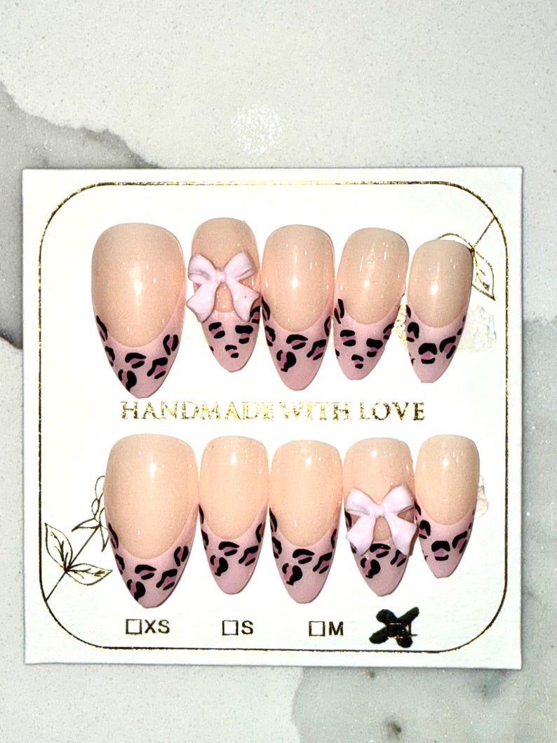 May include: A set of almond-shaped press-on nails. The nails are a nude color with a pink leopard print design on the tips. Some nails feature a small pink bow. The card reads "HANDMADE WITH LOVE".