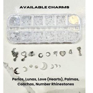 Peut inclure: Un organisateur en plastique transparent rempli de perles et de breloques blanches. En dessous se trouvent divers breloques argent&eacute;es, dont des c&oelig;urs, des lunes et des palmiers. Le texte en haut indique "AVAILABLE CHARMS" et en bas "Perlas, Lunas, Love (Hearts), Palmas, Conchas, Number Rhinestones."