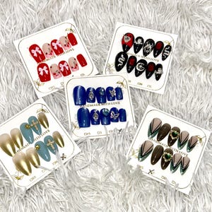 May include: A collection of press-on nail sets with various designs. Sets include red nails with bows, black nails with gems, blue nails with silver accents, gold and blue nails with crosses, and brown nails with green accents.