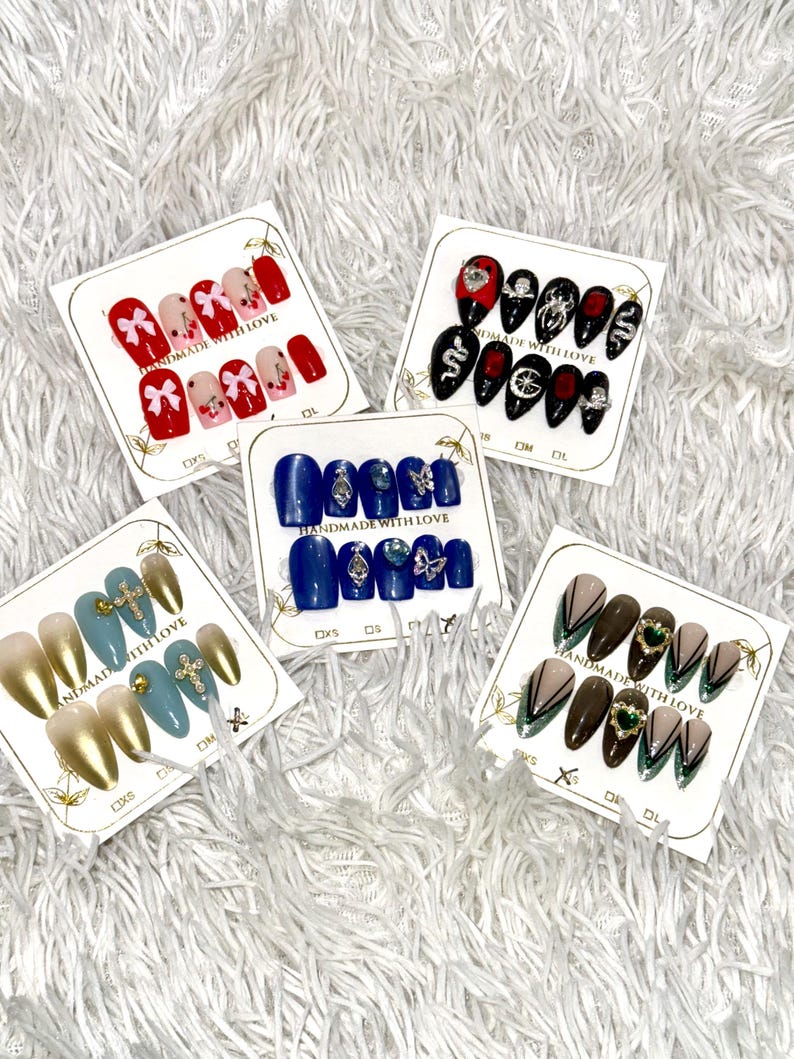 May include: A collection of press-on nail sets with various designs. Sets include red nails with bows, black nails with gems, blue nails with silver accents, gold and blue nails with crosses, and brown nails with green accents.