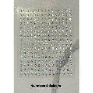 Peut inclure: Feuille transparente d'autocollants num&eacute;rot&eacute;s avec des chiffres m&eacute;talliques argent&eacute;s. Les chiffres de 0 &agrave; 9 sont dispos&eacute;s en rang&eacute;es et en colonnes. Le texte "Number Stickers" est imprim&eacute; en bas de la feuille.