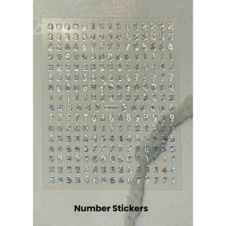 Peut inclure: Feuille transparente d'autocollants num&eacute;rot&eacute;s avec des chiffres m&eacute;talliques argent&eacute;s. Les chiffres de 0 &agrave; 9 sont dispos&eacute;s en rang&eacute;es et en colonnes. Le texte "Number Stickers" est imprim&eacute; en bas de la feuille.
