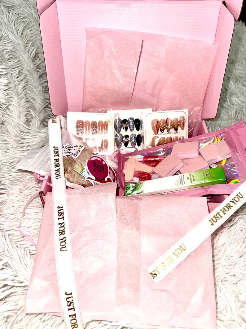 Peut inclure: Une bo&icirc;te cadeau rose remplie de divers accessoires pour nail art, notamment des ensembles de faux ongles, des limes &agrave; ongles et de l'huile &agrave; cuticules. La bo&icirc;te est doubl&eacute;e de papier de soie rose et contient des rubans avec l'inscription dor&eacute;e "JUST FOR YOU".