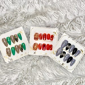 May include: Three sets of false nails in various designs. One set features green and gold glitter nails, another has red and gold accents, and the third has silver glitter and black nails. Each set is displayed on a white card.