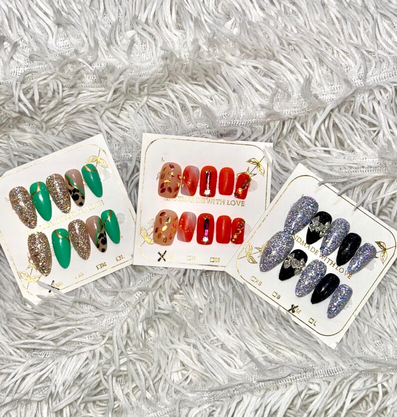May include: Three sets of false nails in various designs. One set features green and gold glitter nails, another has red and gold accents, and the third has silver glitter and black nails. Each set is displayed on a white card.