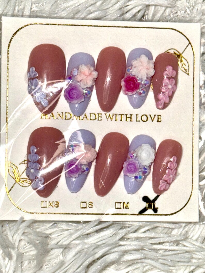 May include: A set of artificial nails in various sizes, featuring a mix of mauve and lavender colors. Some nails are adorned with floral embellishments, including small pink and purple flowers. The packaging reads "HANDMADE WITH LOVE."