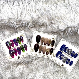 May include: Three sets of false nails on white cards. One set features a mix of pink, purple, and green nails with glitter and gems. Another set has brown, nude, and gold nails with heart and bow accents. The third set has blue and gray nails with silver embellishments.
