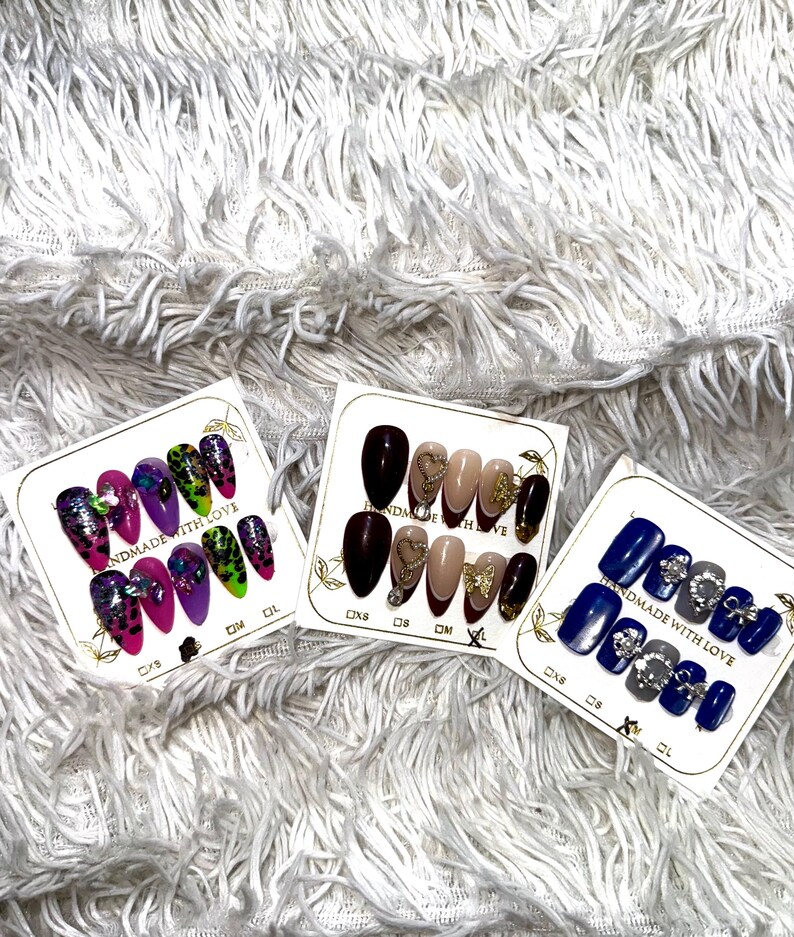 May include: Three sets of false nails on white cards. One set features a mix of pink, purple, and green nails with glitter and gems. Another set has brown, nude, and gold nails with heart and bow accents. The third set has blue and gray nails with silver embellishments.