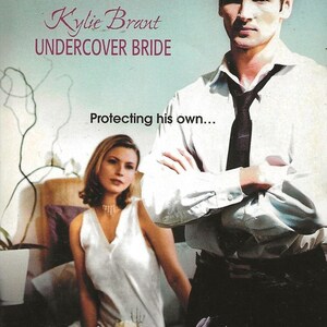May include: A book cover with a couple on the front. The man is wearing a white shirt and a dark tie. The woman is wearing a white sleeveless dress. The title of the book is "Kylie Brant UNDERCOVER BRIDE" and the tagline is "Protecting his own... A Year of Loving Dangerously".