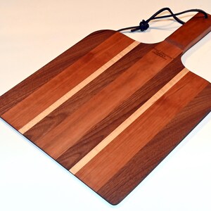May include: A rectangular wooden pizza cutting board with a handle. The board is made of walnut and cherry wood with maple strips. The board has a black leather strap attached to the handle.