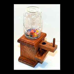 May include: A wooden candy dispenser with a glass jar on top. The dispenser is made of cherry wood and has a crank on the side. The jar is filled with colorful candies.