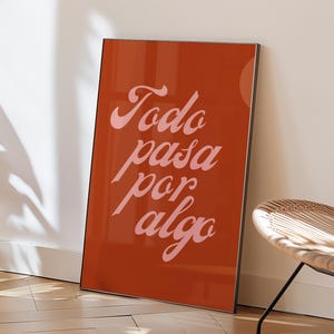Op de afbeelding: Een oranje ingelijste kunstprint met de tekst "Todo pasa por algo" in roze cursief schrift. Het kunstwerk leunt tegen een witte muur, met een houten vloer en een rotan stoel op de achtergrond.