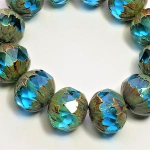 May include: A strand of teal blue glass beads with a textured, aged, metallic finish. The beads are faceted and have a unique, iridescent quality.