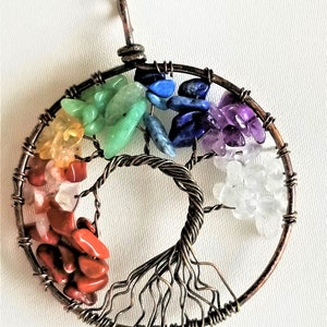 May include: A handmade pendant with a wire tree design, embellished with colourful gemstones. The circular frame is decorated with a spectrum of stones: red, orange, yellow, green, blue, and purple, representing the chakras. The tree's trunk and branches are intricately crafted.