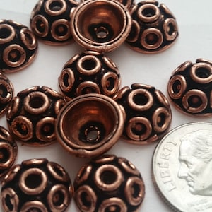 Puede incluir: Un grupo de 12 cuentas de metal de color cobre en forma de cúpula con un patrón circular. Las cuentas son aproximadamente del tamaño de una moneda de diez céntimos de euro.