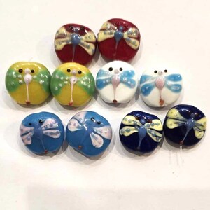 May include: Assortment of ten glass beads with dragonfly designs. The beads are in various colors, including red, yellow, green, blue, and white.