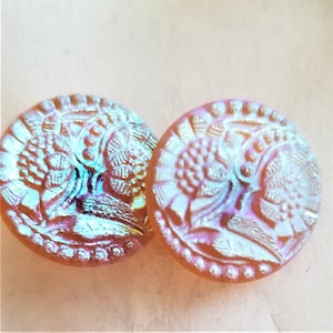 Puede incluir: Dos botones de vidrio iridiscente con un diseño floral. Los botones son de color rosa pálido con un brillo iridiscente blanco.