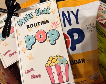 May include: A gift set featuring a bag of cheddar popcorn and a decorative tag. The tag reads "Make that ROUTINE POP" with a cartoon popcorn graphic and the text "Love, Clara". The bag is white and yellow.