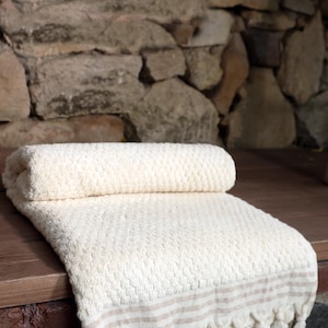 May include: A white waffle-textured bath towel with a brown and white striped border and fringe. The towel is rolled up and placed on a wooden surface.