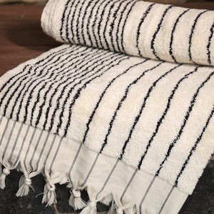 May include: A white bath towel with black stripes. The towel has a fringe border and is folded in half.