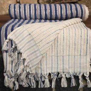 May include: A white and blue striped Turkish towel with fringe. The towel is folded in half and has a blue and gray striped towel on top of it.