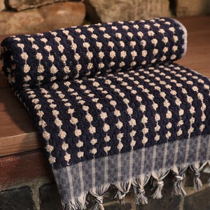 May include: A navy blue and cream-colored patterned towel with a textured design. The towel is folded and rolled, with a fringe detail at the bottom. The towel is made of a soft material.