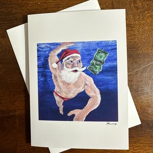 May include: A white greeting card featuring an illustration of Santa Claus in a red hat and speedo, reaching for a dollar bill. The background is blue, and the card is paired with a white envelope. The artwork is signed.