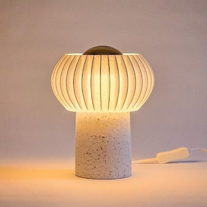 Könnte beinhalten: Eine weiße Tischlampe mit einem weißen, gerippten Lampenschirm und einem weißen, strukturierten Sockel. Die Lampe ist eingeschaltet und das Licht scheint durch den Lampenschirm.