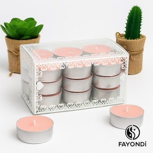Puede incluir: Una caja de plástico transparente llena de 12 velas de té de color rosa. Las velas están en un soporte plateado y la caja tiene un diseño de corazón blanco. La caja está sobre una superficie blanca.