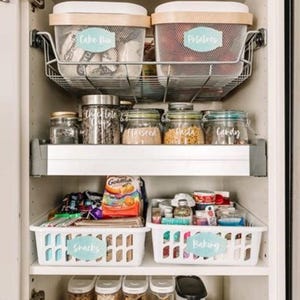Pode incluir: Despensa organizada com vários recipientes e caixas rotuladas. Inclui caixas de plástico branco para biscoitos, bolachas e sopa. Cestos de arame contêm mistura para bolo e batatas. Frascos contêm sementes, massa e doces.
