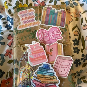 Puede incluir: Un paquete de papel marrón envuelto con cuerda y decorado con pegatinas coloridas. Las pegatinas presentan ilustraciones y texto con temática de libros, incluyendo "Get in, we're going book shopping", "Hot girls read books", "Enemies to lovers", "Fake dating", "Forced proximity", "Men written by woman", y "If I had a dollar for every fictional character I caught feelings for, I could afford the therapy, I so obviously need".