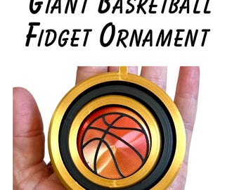 May include: A round fidget ornament featuring a central orange basketball graphic with black lines, encased in a black ring and a larger gold outer frame. The text GIANT BASKETBALL FIDGET ORNAMENT is printed in black above the circular object.