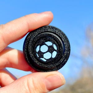 Puede incluir: Objeto circular negro con una superficie texturizada en relieve. El centro del objeto tiene un diseño recortado de un balón de fútbol.