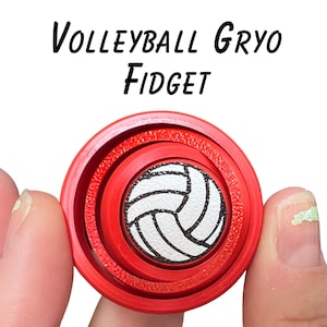 Puede incluir: Un juguete fidget con temática de voleibol, rojo y blanco. El centro presenta un diseño de voleibol blanco con líneas negras, rodeado de anillos rojos concéntricos. El texto "Volleyball Gryo Fidget" está arriba.