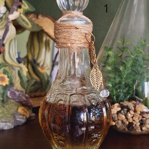 Op de afbeelding: Een heldere glazen fles met een bruine vloeistof erin. De fles heeft een kurken stop en is versierd met touw en gouden bladcharms. De fles staat op een houten oppervlak.